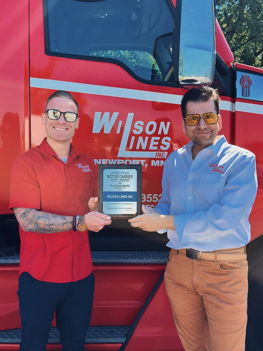Two Wilson Lines team members holding a safety award beside a red truck.