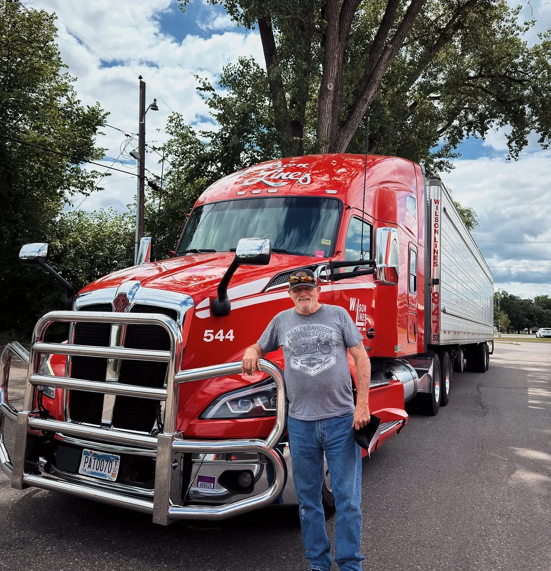 Wilson Lines driver beside red tractor-trailer 544 beneath the trees.
